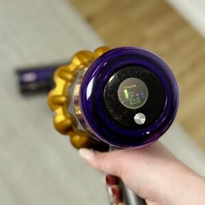 Das Display des Dyson V15 Detect zeigt den aktiven Eco-Modus. Eine Hand hält das Gerät über einem Teppichboden.
