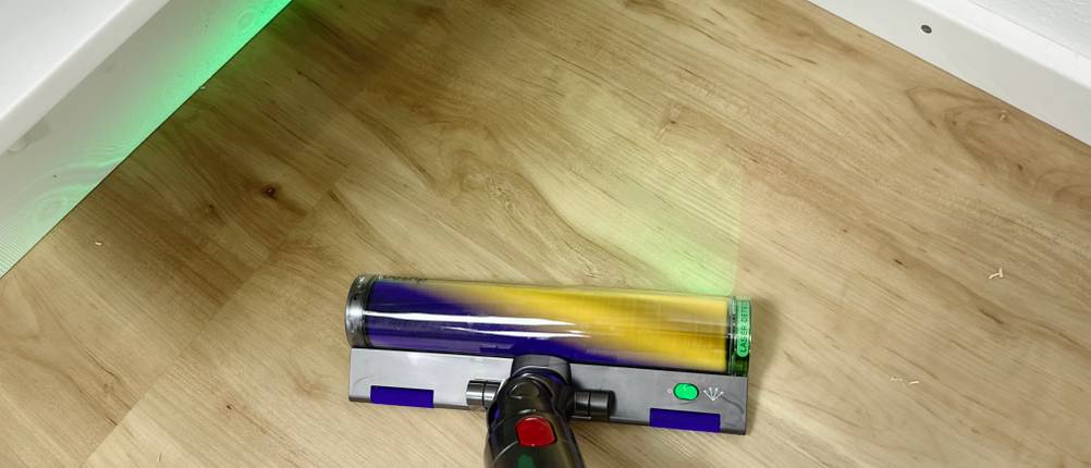 Die Laser Slim Fluffy-Bürste des Dyson V15 Detect leuchtet grünes Licht auf den Boden. Der Sauger wird unter einem Regal verwendet.