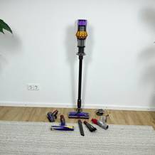 Der Dyson V15 Detect mit aufgestecktem Rohr und allen Zubehörteilen auf dem Boden verteilt. Das Gerät steht senkrecht an der Wand.