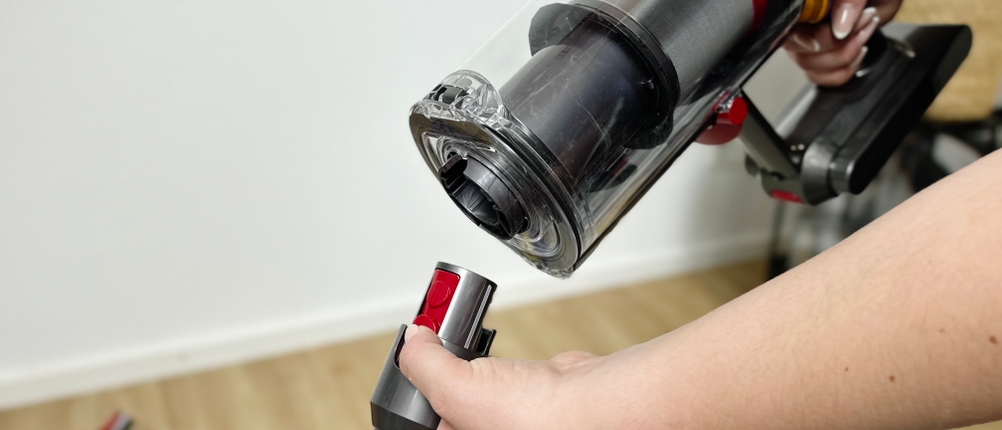 Das Bild zeigt, wie das Verlängerungsrohr des Dyson V15 Detect mit dem Hauptgerät verbunden wird. Eine Hand drückt den roten Verriegelungsknopf am Rohr.