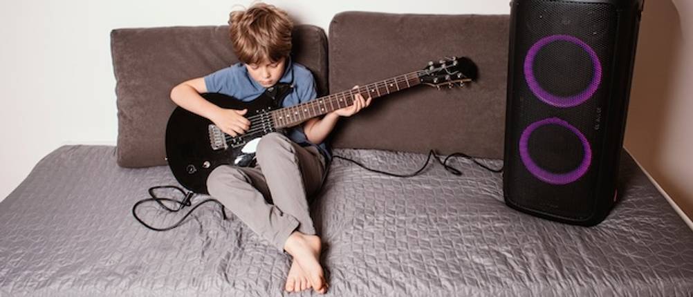 E-Gitarre für Kinder im Test: Ein Junge sitzt auf einem großen, grauen Sofa neben einer großen Verstärkerbox und spielt auf einer schwarzen E-Gitarre.