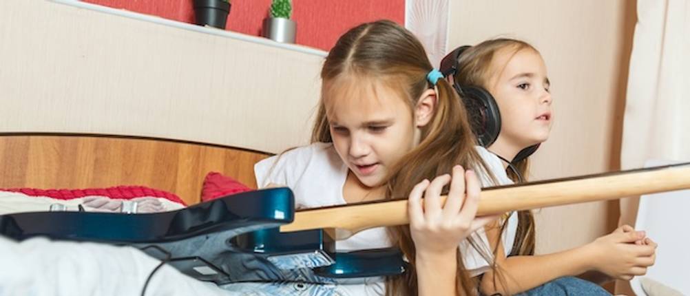 Ein Mädchen spielt auf einer getesteten E-Gitarre für Kinder. Dahinter sitzt ein weiteres Mädchen mit Kopfhörern.