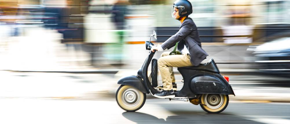 Ein Mann sitzt mit einem schwarzen Helm auf einem schwarzen Roller und fährt auf einer Straße.