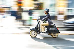 Test der E-Scooter-Versicherungen: Ein Mann sitzt mit einem schwarzen Helm auf einem schwarzen Roller und fährt auf einer Straße.