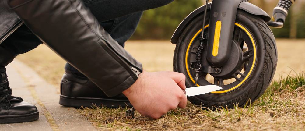 E-Scooter-Versicherungen im Test: Ein Mann hält ein Messer an den Reifen eines E-Scooters.