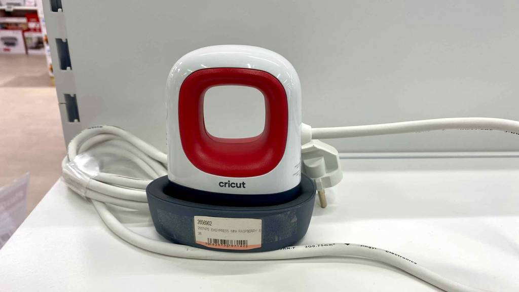 Nahaufnahme von vorn einer Transferpresse im Test: weiß-rotes Modell Easypress Mini der Marke Cricut inklusive schwarzer Basis und weißem Kabel befindet sich auf einem weißen Verkaufsregal.