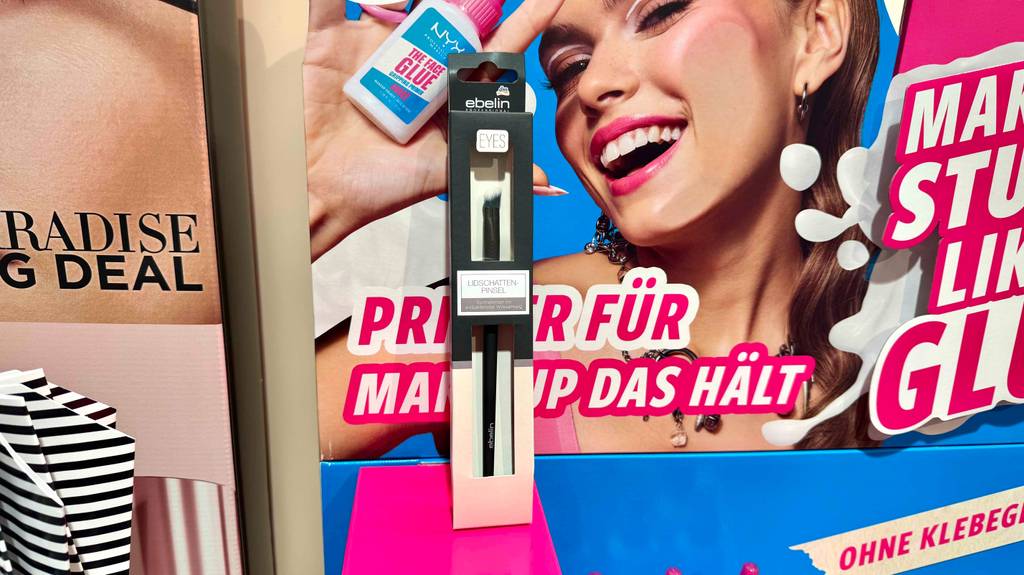 Ein verpackter Lidschattenpinsel steht zum testen vor einem Werbeposter.