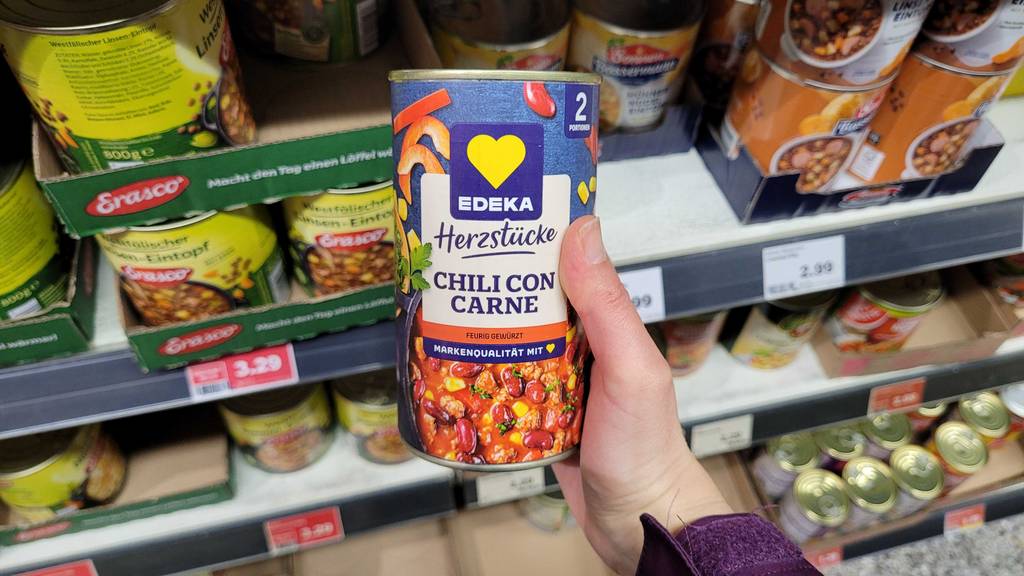 Eine Hand hält eine Dose Chili con Carne im Test, die zwei Portionen enthält, in einem Verkaufsbereich.