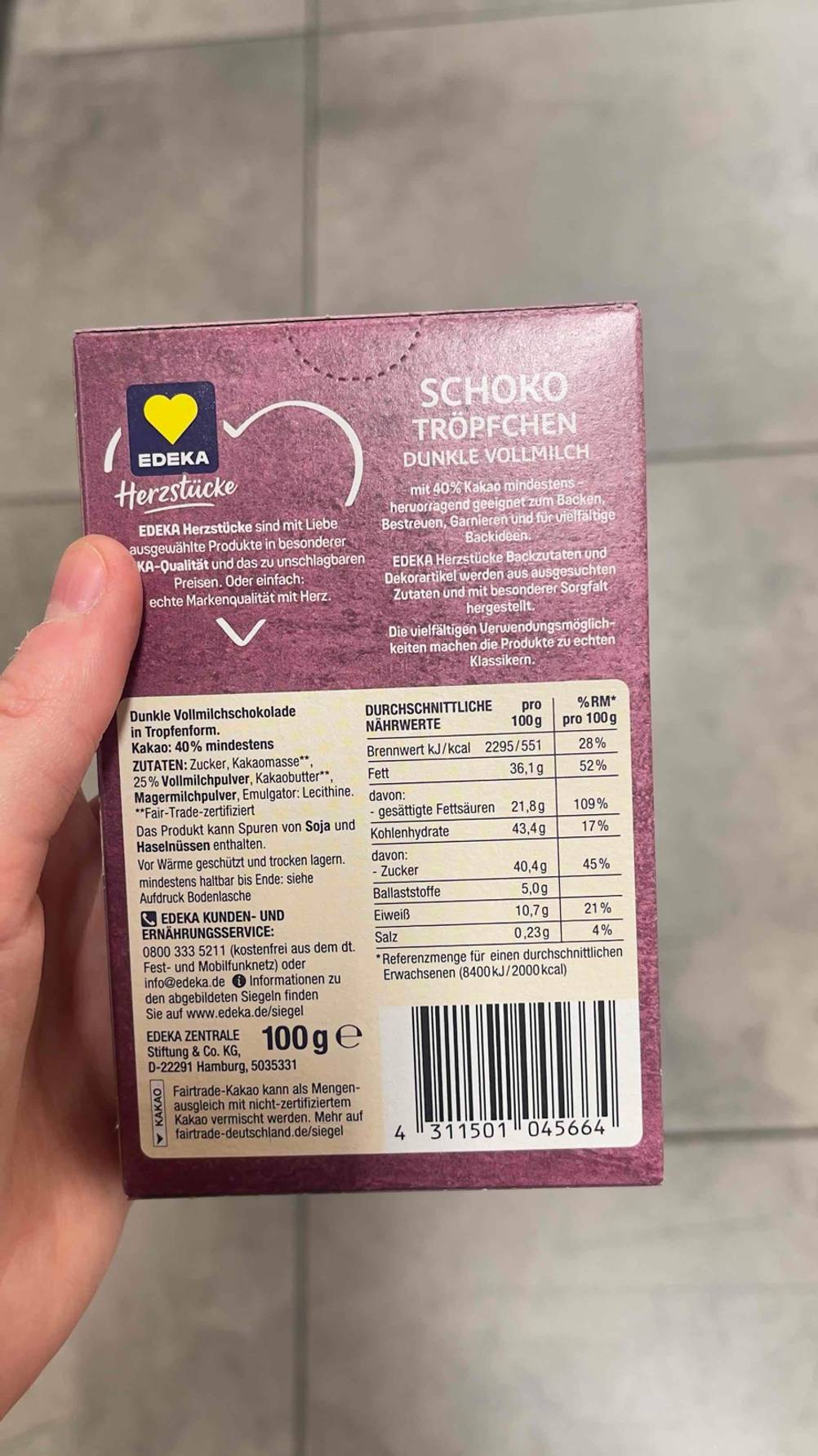 Getestet: Die Schokodrops der Marke EDEKA in einer 100-g-Packung, inklusive Produktangaben, die sich in einer Hand über einem grauen Fliesenboden befindet.