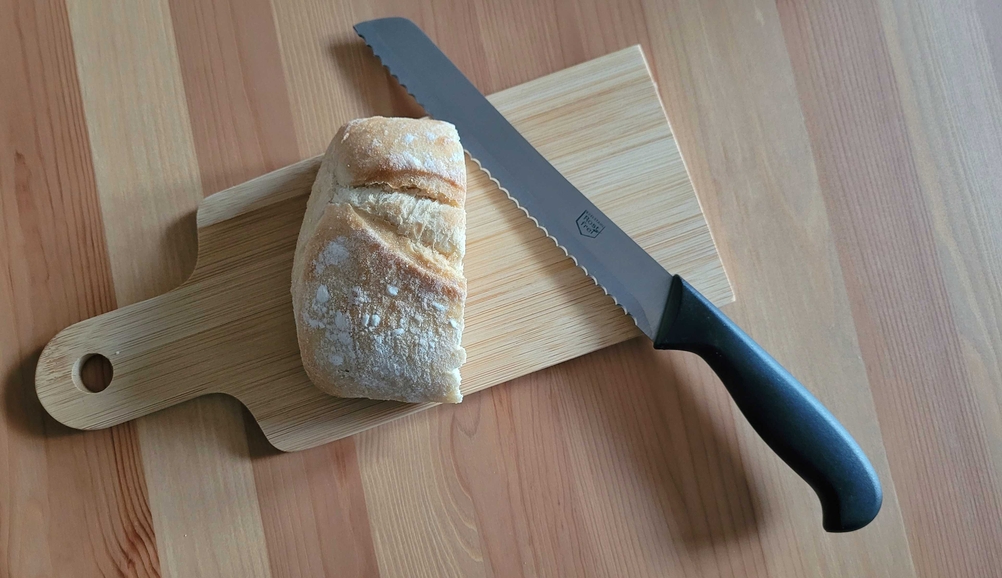 Brotmesser im Test: Auf einem Holzbrett liegen ein Brotmesser und ein Stück Weißbrot nebeneinander.
