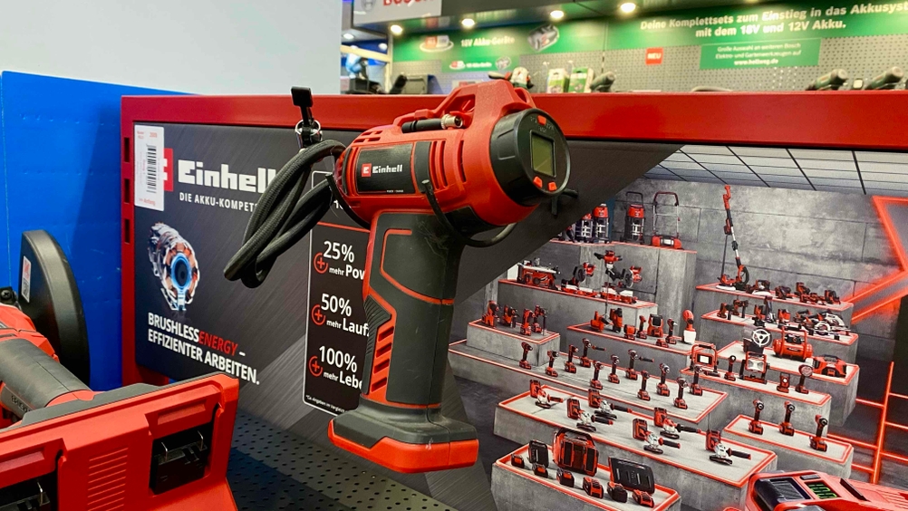 Eine getestete Akku-Luftpumpe hängt an einem Verkaufsregal, im Hintergrund ein Plakat der Marke Einhell.