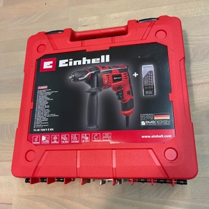 Transportkoffer der Einhell-TC-ID-720/1-E-Schlagbohrmaschine auf einem Holztisch liegend. Der Koffer zeigt die Maschine und ein Bohrerset auf der Verpackung.