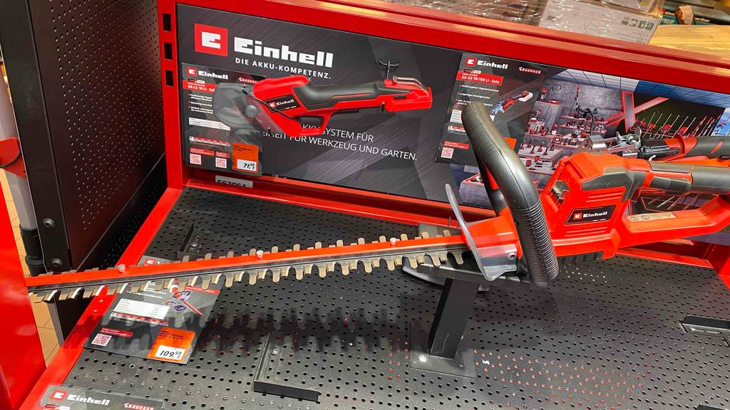 Seitenansicht einer getesteten Einhell-Heckenschere im roten Design, mit langem Sägeblatt auf einem Verkaufsregal vor dem Karton eines Einhell-Gartengeräts.