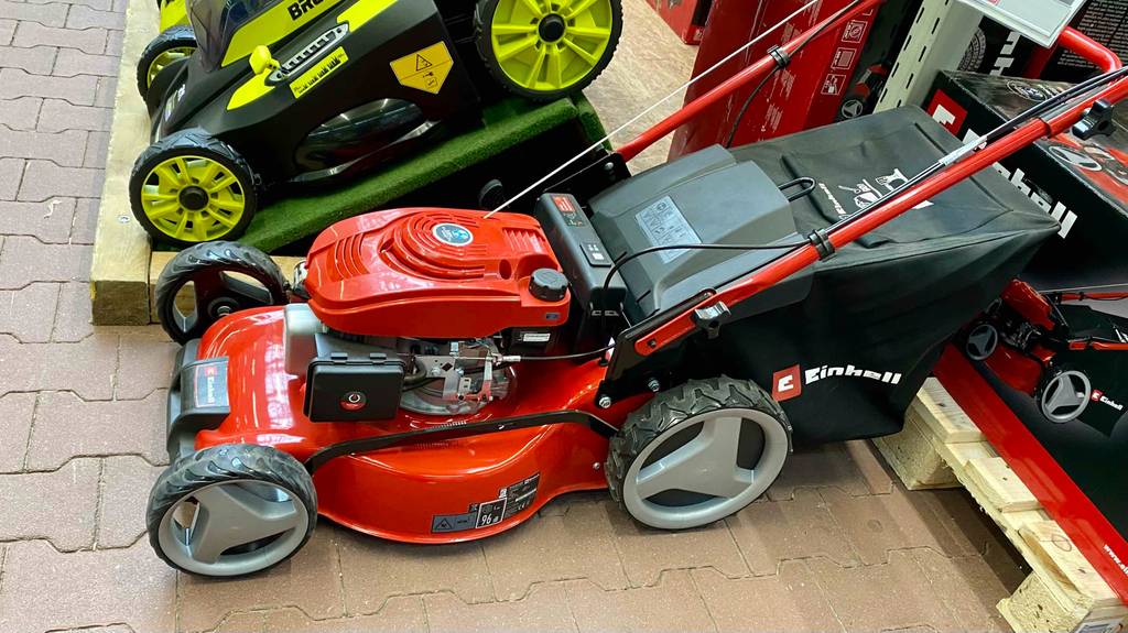 Seitenansicht von schräg oben auf einen roten Einhell Benzin-Rasenmäher im Test, inklusive schwarzem Fangkorb und verlängerbarem Griff in einem Verkaufsbereich mit weiteren Rasenmähern.