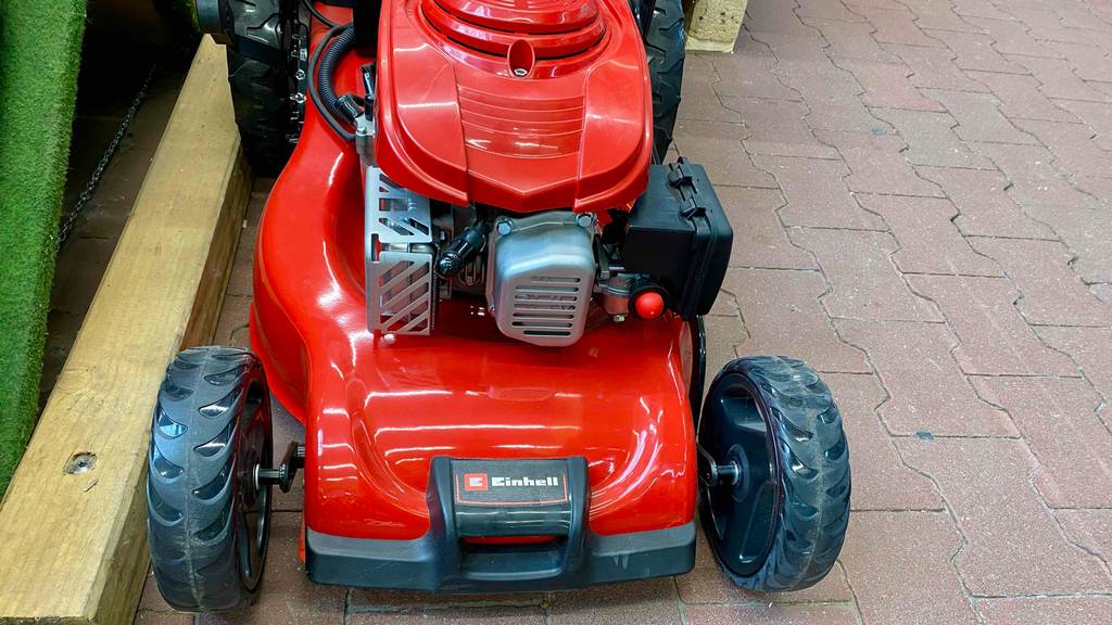 Detailansicht von vorn des Motorblocks inklusive Elektrostarter eines roten Einhell Benzin-Rasenmähers im Test, der sich auf roten Steinfliesen in einem Verkaufsbereich befindet.
