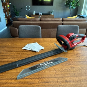 Die Einhell GE-EH 7067 liegt mit Schutz über dem Scherblatt neben dem Schnittgutsammler und den Anleitungen auf einem Holztisch.