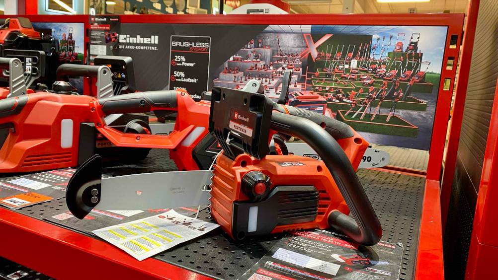 Getestet: Eine Kettensäge von der Seite auf einer dunklen Fläche stehend, im Hintergrund ein Plakat der Markte Einhell und andere Geräte.