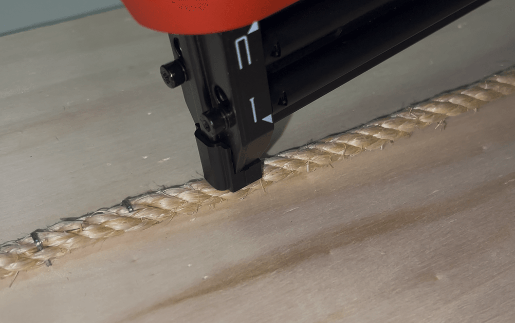 Der Einhell TE-CN 18 Li Akku-Tacker fixiert eine dicke Juteschnur mit Heftklammern auf einer Holzplatte. Die Klammern liegen gleichmäßig und präzise an der Kordel an.