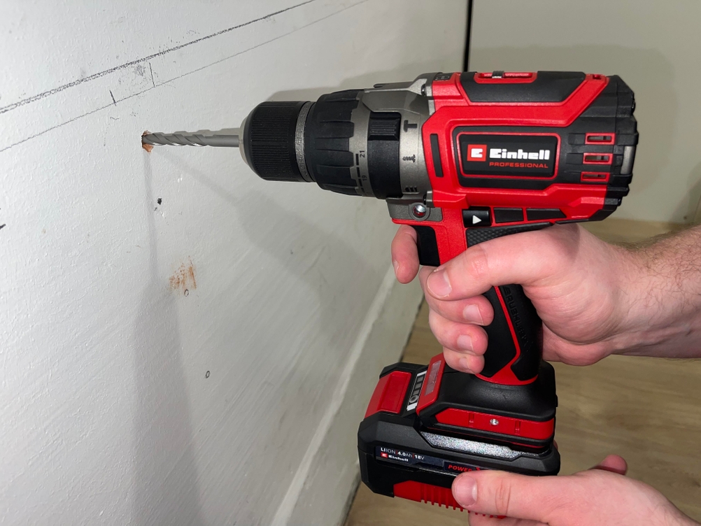 Auf dem Bild ist der Einhell Professional TP-CD 18/70 Li BL im Einsatz auf einer Werkbank im Freien zu sehen. Der Bohrer ist in Holz oder beschichtetes Plattenmaterial eingedrungen.