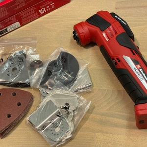 Das Bild zeigt den Einhell Varrito samt Zubehörteilen in der Unboxing-Szene. Verschiedene Sägeblätter und Schleifpapiere liegen neben dem Gerät.