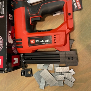 Der Einhell TE-CN 18 Li Akku-Tacker liegt mit Zubehör, Anleitung und mehreren Heftklammer-Typen neben der geöffneten Verpackung. 