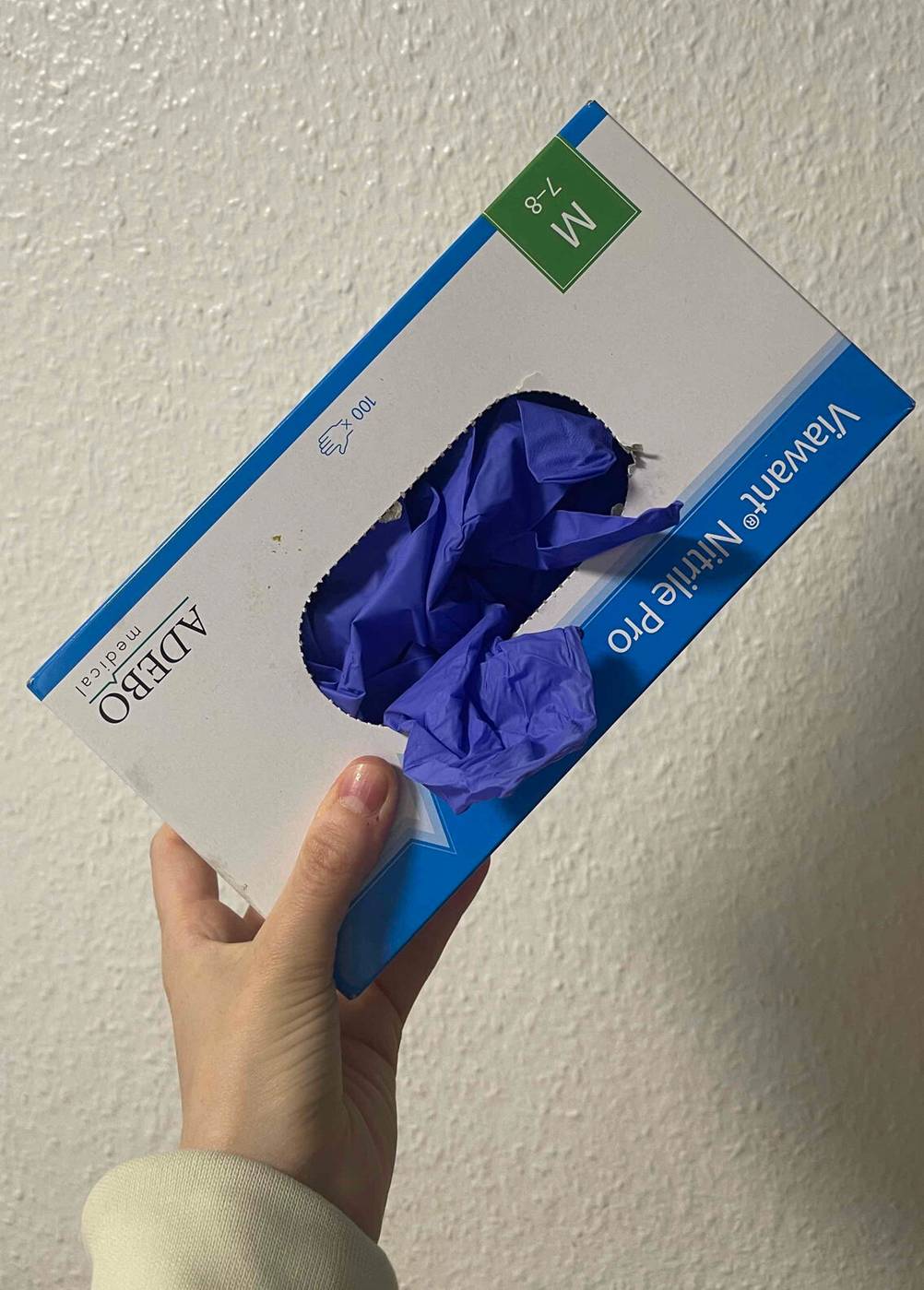 Eine Hand hält einen Spender-Karton mit den getesteten Einmalhandschuhen (100 Stück) in Violett vor einer weißen Wand.
