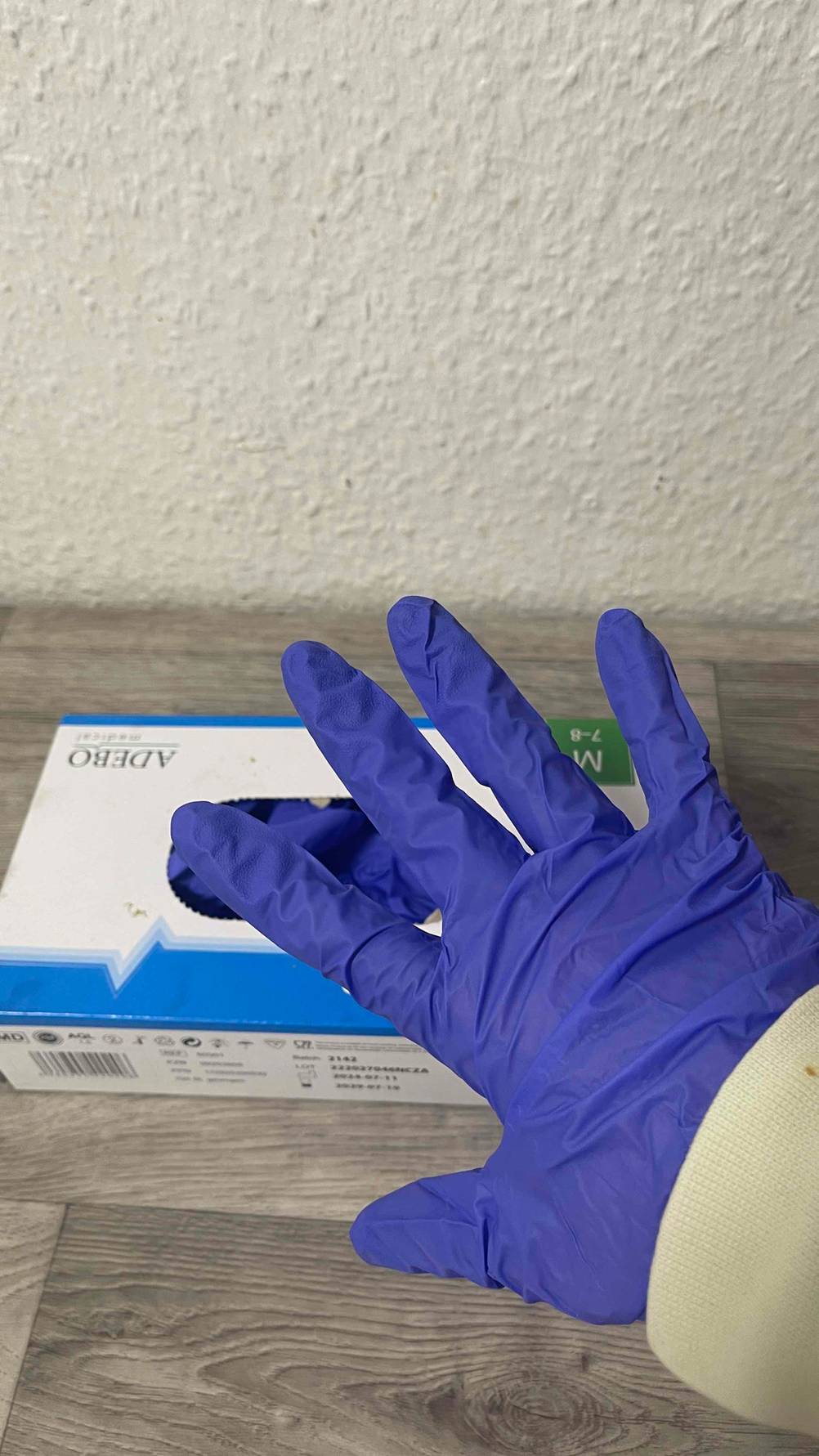 Test: Die Einmalhandschuhe (100 Stück) in Violett in einem Spender-Karton, der auf einer Holzfläche steht. Darüber eine behandschuhte Hand.