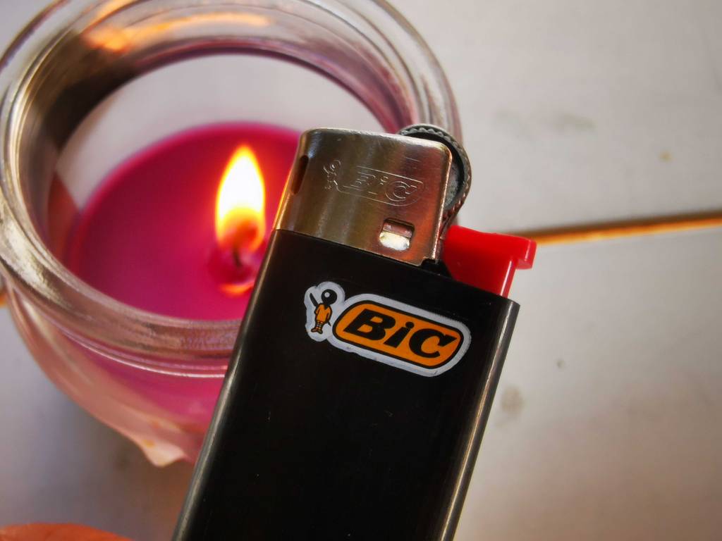 Nahaufnahme des schwarzen, getesteten Einwegfeuerzeuges der Marke Bic über einer brennenden Kerze im Glas.
