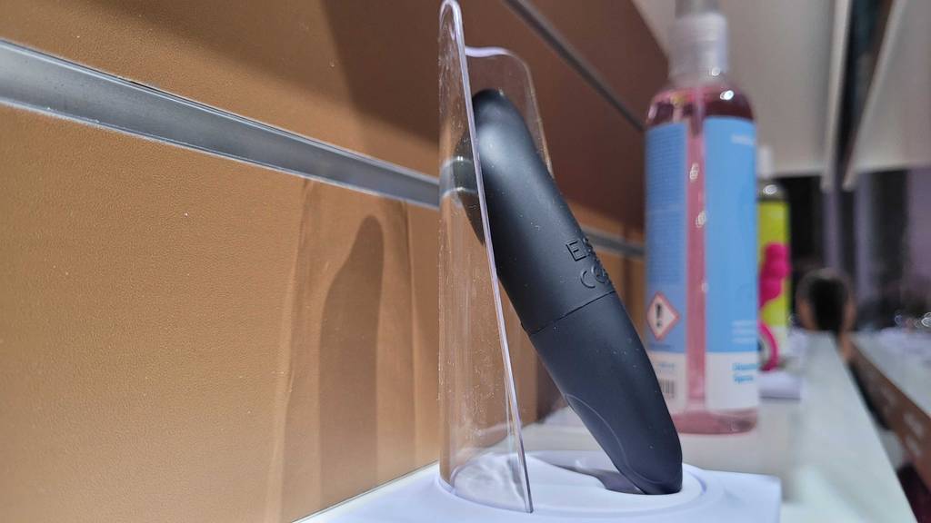 Squirting-Dildo getestet: seitliche Nahaufnahme eines schwarzen Dildos in einem Plexiglas-Ständer auf einem Ausstellungsregal mit anderen Produkten.