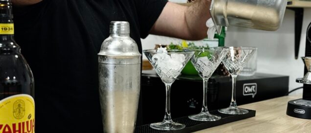 Ein Barkeeper befüllt Cocktailgläser mit Eiswürfeln aus einem Metallbehälter. Im Vordergrund steht eine Flasche Kaffeelikör.