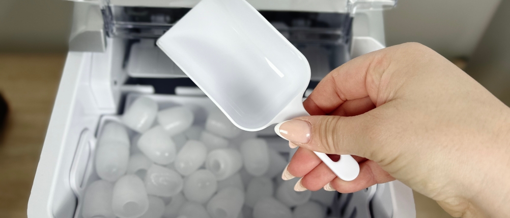 Eine Hand hält eine weiße Eisschaufel über den gefüllten Eisbehälter der Conopu ICM1227. Der Deckel ist geöffnet.