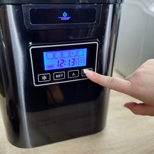 Eine Hand drückt eine Taste am beleuchteten Bedienfeld der Kesser 14812 Eiswürfelmaschine. Das Display zeigt Uhrzeit und Symbole.