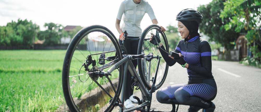 Zwei Radfahrerinnen reparieren einen platten Rennradreifen auf einer Landstraße im Grünen.