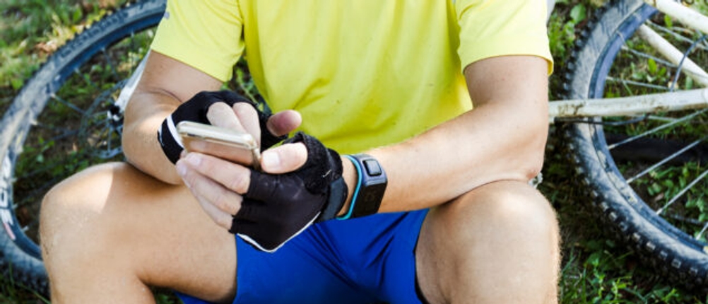 Ein Radfahrer in Radkleidung sitzt am Waldrand und lädt sein Smartphone mit einer Powerbank auf.
