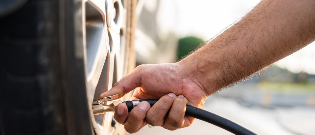 Eine Person nutzt eine elektrische Luftpumpe, um einen Autoreifen über das Ventil mit Luft zu befüllen. Der Schlauch ist am Ventil angeschlossen, die Hand hält den Adapter fest.