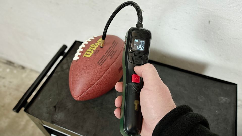 Eine Hand hält die Bosch Easypump 603947000, die mit einem Ball verbunden ist. Das Display zeigt den aktuellen und voreingestellten Luftdruck an.