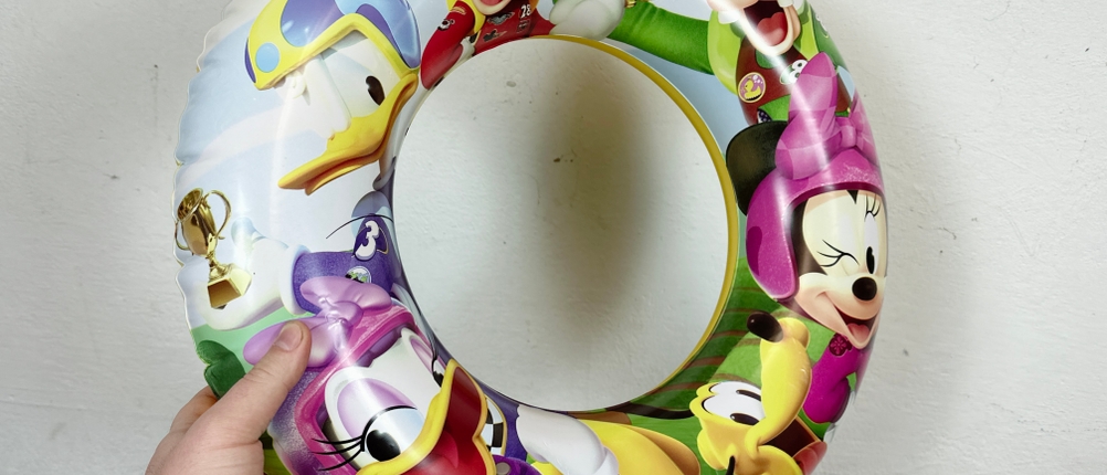 Eine Hand hält einen bunten, aufgepumpten Schwimmreifen mit Disney-Motiven vor einer weißen Wand. Der Reifen ist vollständig mit Luft gefüllt.