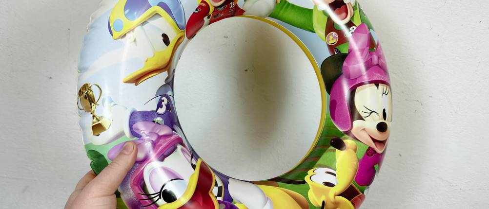 Eine Hand hält einen bunten, aufgepumpten Schwimmreifen mit Disney-Motiven vor einer weißen Wand. Der Reifen ist vollständig mit Luft gefüllt.
