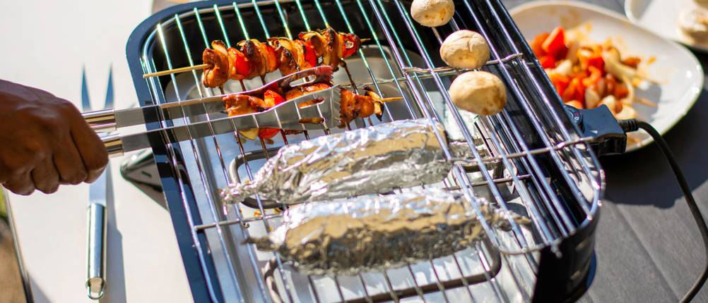 Elektrogrill mit Spießen, Kartoffeln und Folien-Grillgut steht auf einer hellen Ablage neben einem Gartentisch.