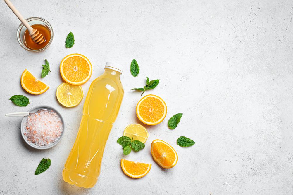 Eine Getränkeflasche mit Inhalt liegt auf einer Unterlage. Darum verteilt liegen Zitronen- und Orangenscheiben sowie Kräuterblätter, ein Schälchen mit Honig und ein Schälchen mit Salz.