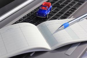 Test von elektronischen Fahrtenbüchern: Ein herkömmliches Fahrtenbuch liegt zusammen mit einem Kugelschreiber und einem Spielzeugauto auf einer Tastatur.