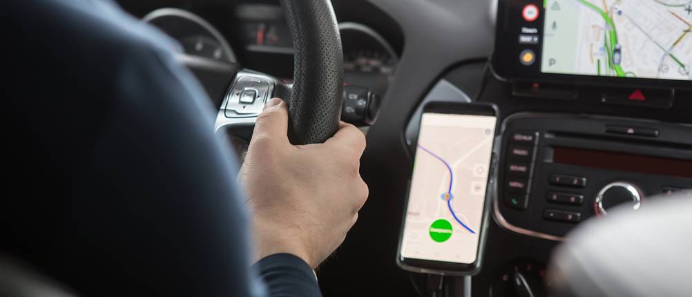 Elektronische Fahrtenbücher im Test: Ein Mann sitzt am Steuer eines Autos, neben ihm befindet sich ein Navigationsgerät und ein Smartphone mit einer Navigations-App.