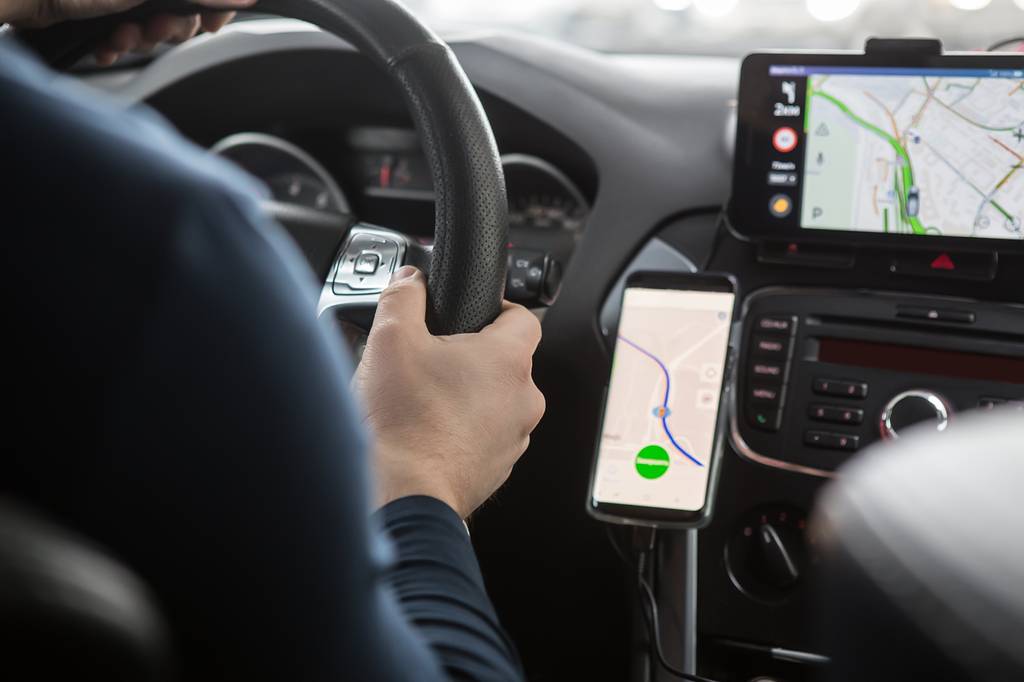 Ein Mann sitzt am Steuer eines Autos, neben ihm befindet sich ein Navigationsgerät und ein Smartphone mit einer Navigations-App.