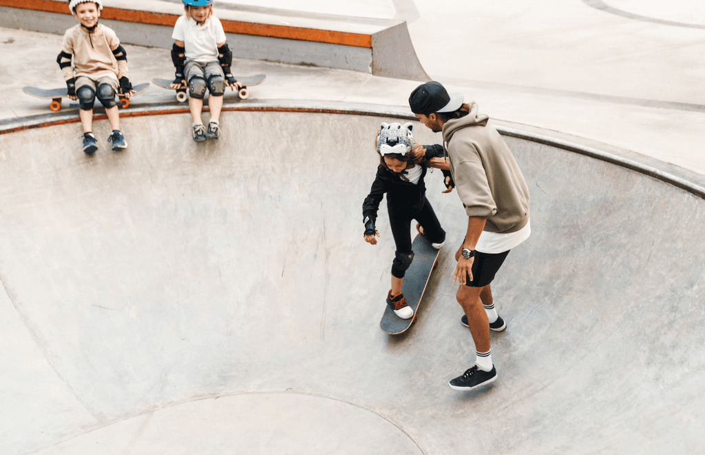 Ellenbogenschoner-Test: In einem Skatepool skatet ein kleines Mädchen mit Hilfe ihres Vaters. An der Rampe sitzen zwei weitere Kinder mit Skateboard. 