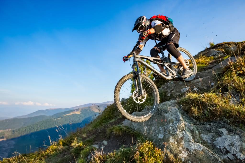 Ellebogenschoner-Test: Ein Mann fährt mit einem Enduro-Mountainbike auf einem felsigen Trail. Im Hintergrund sind Berge und ein blauer Himmel zu sehen. 