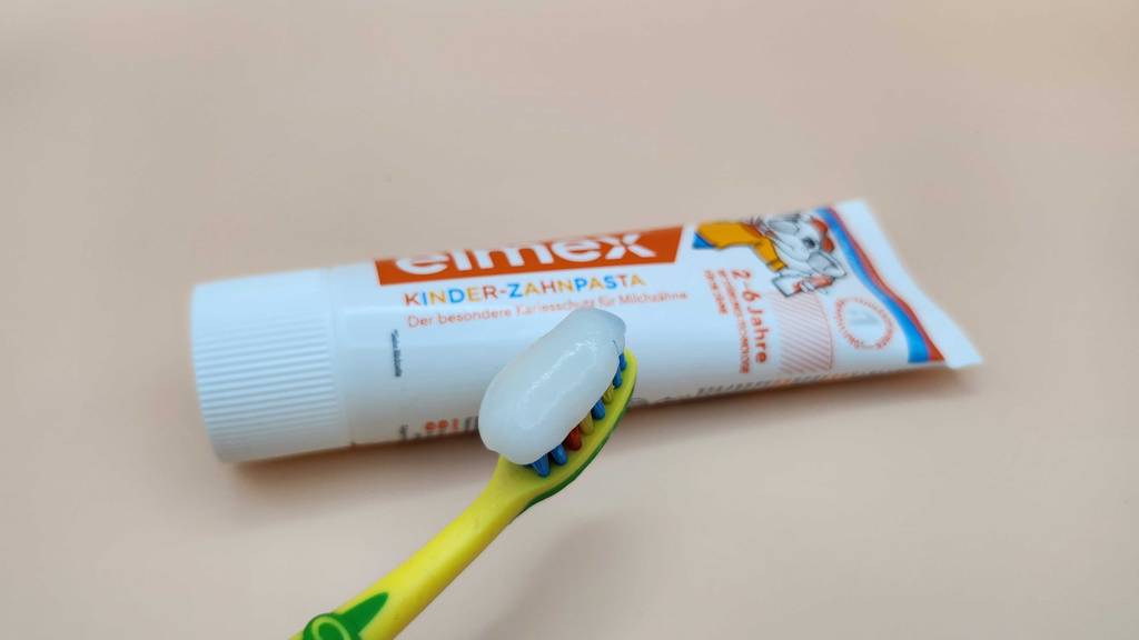 Auf einer Zahnbürste ist ein Klecks Elmex Zahnpasta zum testen, die Tube im Hintergrund.