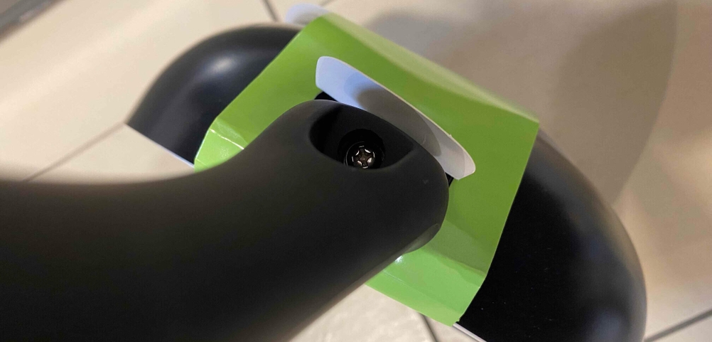 Beschichtete Pfannen im Test: Nahaufnahme des angeschraubten Griffs einer original verpackten schwarzen Pfanne vor beigefarbenen Fliesen.