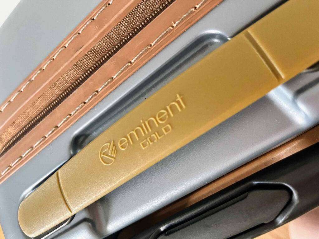 Nahaufnahme des ausziehbaren schwarzen Handgriffs, des goldfarbenen Handgriffs und des Reißverschlusses eines getesteten leichten Koffers der Marke Eminent Gold.