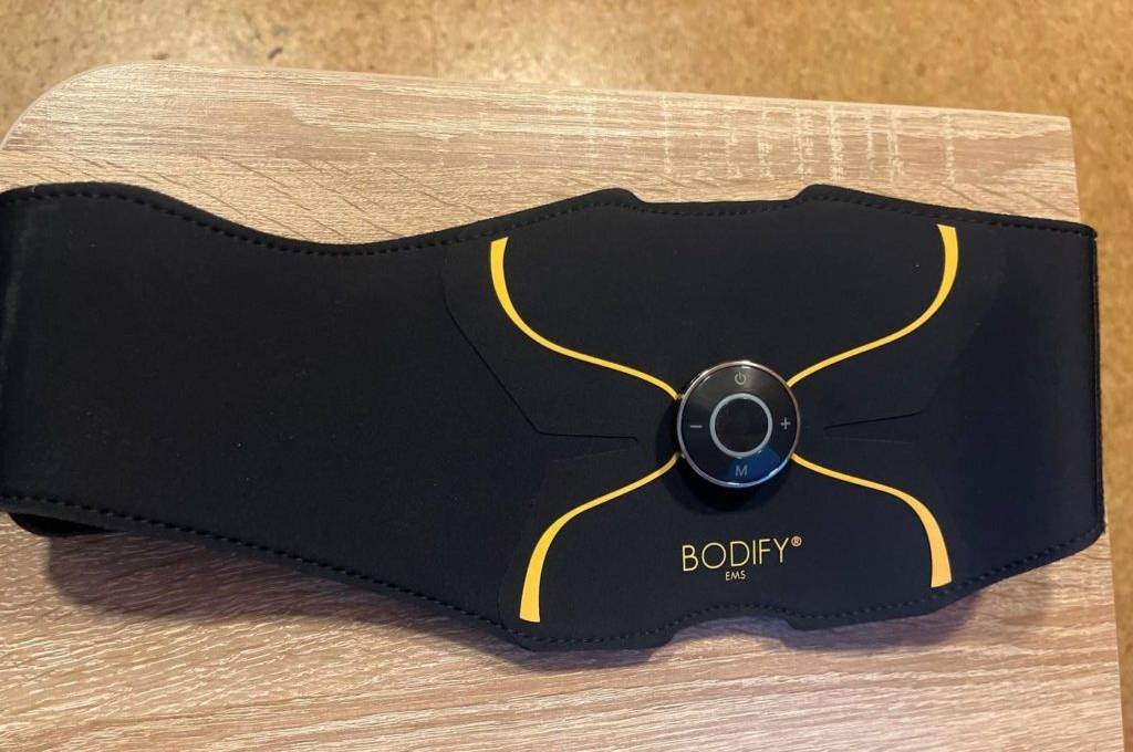 Ein schwarzer Bodify Ems Bauchmuskelgürtel im Test mit gelben Verzierungen liegt mit dem Einstellknopf nach oben auf einer hellen Holzfläche.