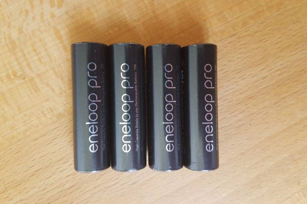 Vier schwarze, getestete AA-Akkus des Typs eneloop pro liegen auf einem hellen Holzuntergrund.